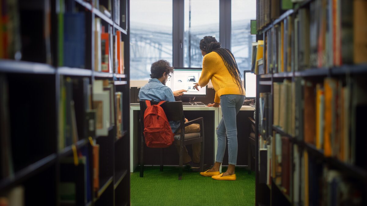 Two students standing and sitting at a desk with a computer in a library, surrounded by bookshelves.