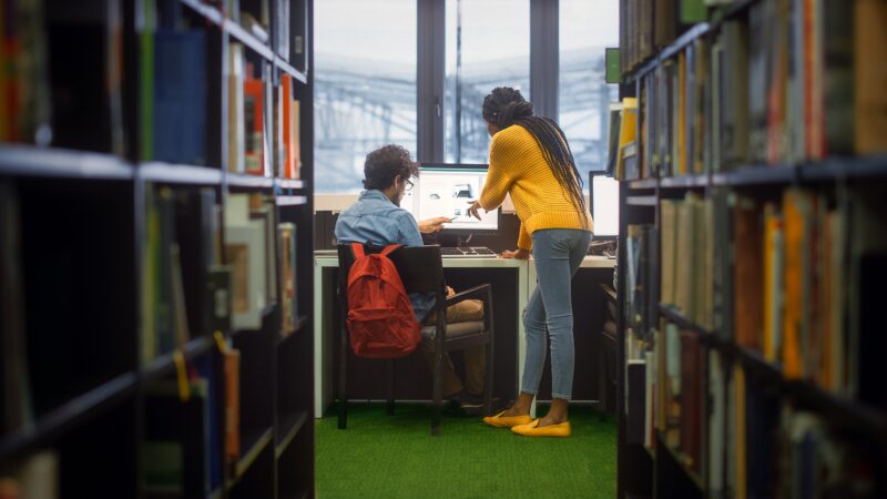 Two students standing and sitting at a desk with a computer in a library, surrounded by bookshelves.
