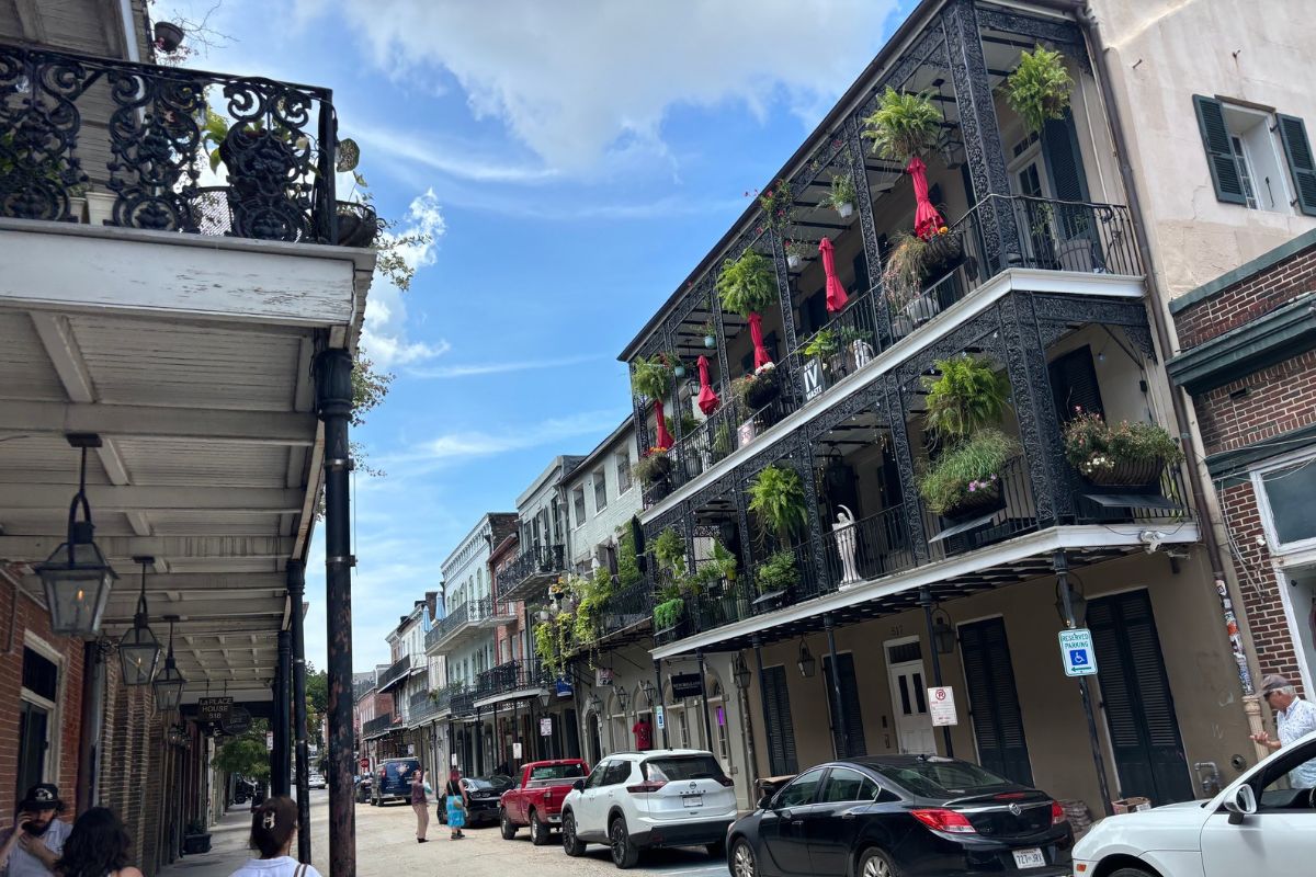 A street view in New Orleans featuring historic buildings with black iron balconies decorated with hanging plants and red umbrellas.