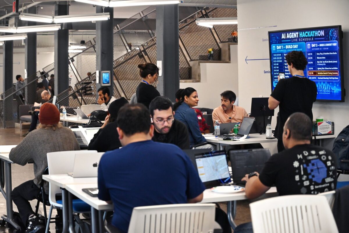 People working at laptops in a collaborative office space with an AI Agent Hackathon schedule displayed on a wall-mounted monitor.