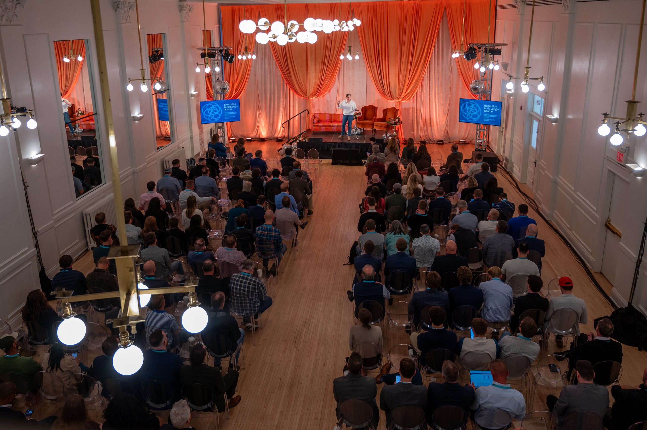 A speaker stands on a stage before an audience in a large, high-ceilinged hall with orange curtains.