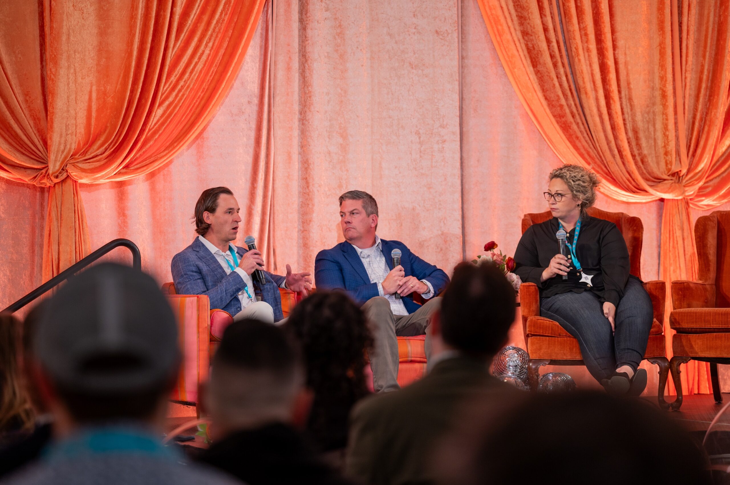 Two men and a woman sit in armchairs on a stage holding microphones, participating in a panel discussion.