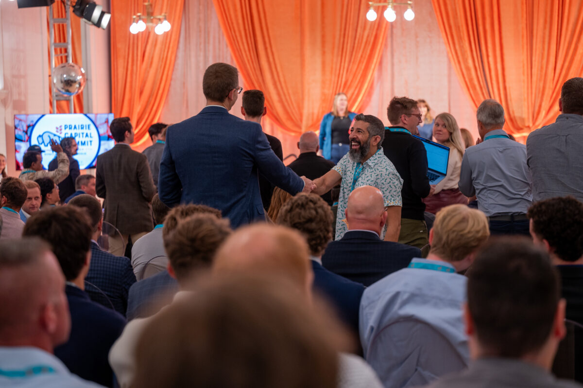 Two men shake hands in the center of a crowded conference room in front of a stage with orange curtains.