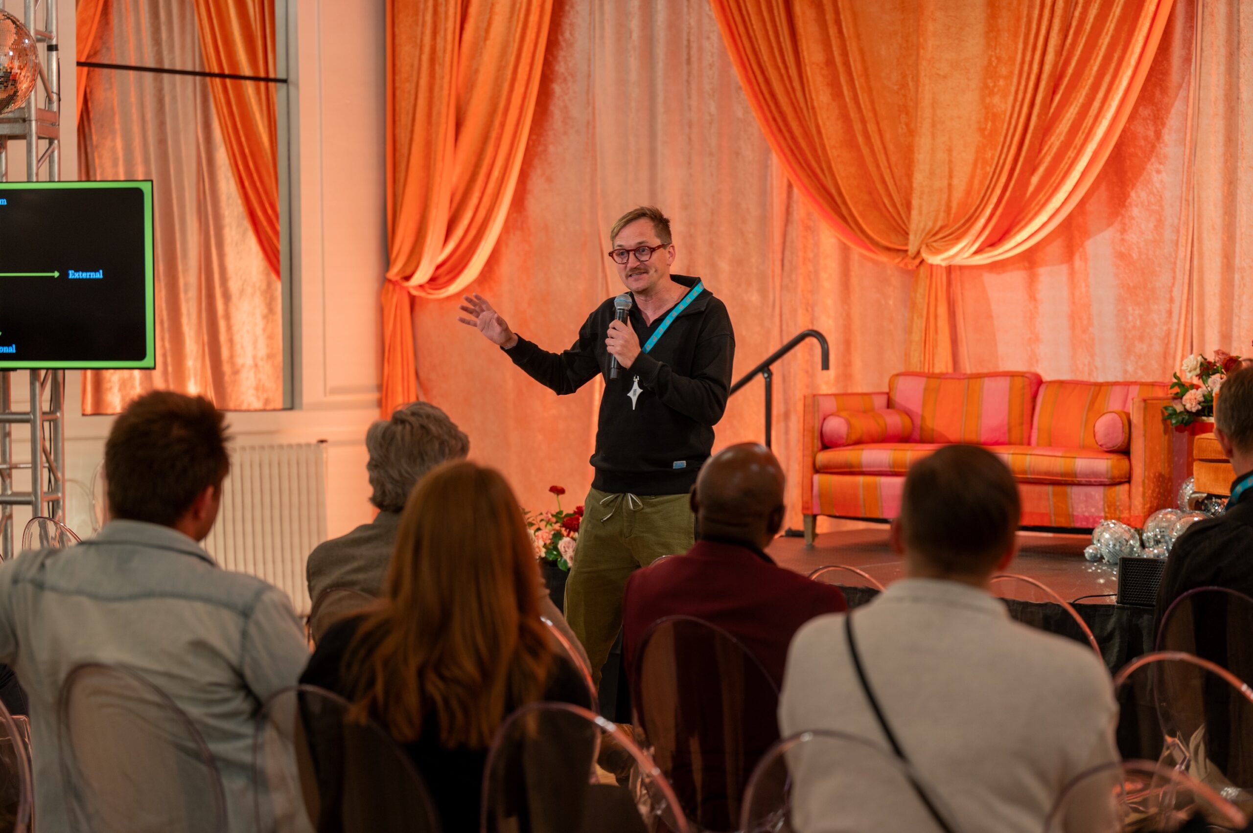 A man in a black sweatshirt and glasses gestures while speaking into a microphone on a stage before an audience.