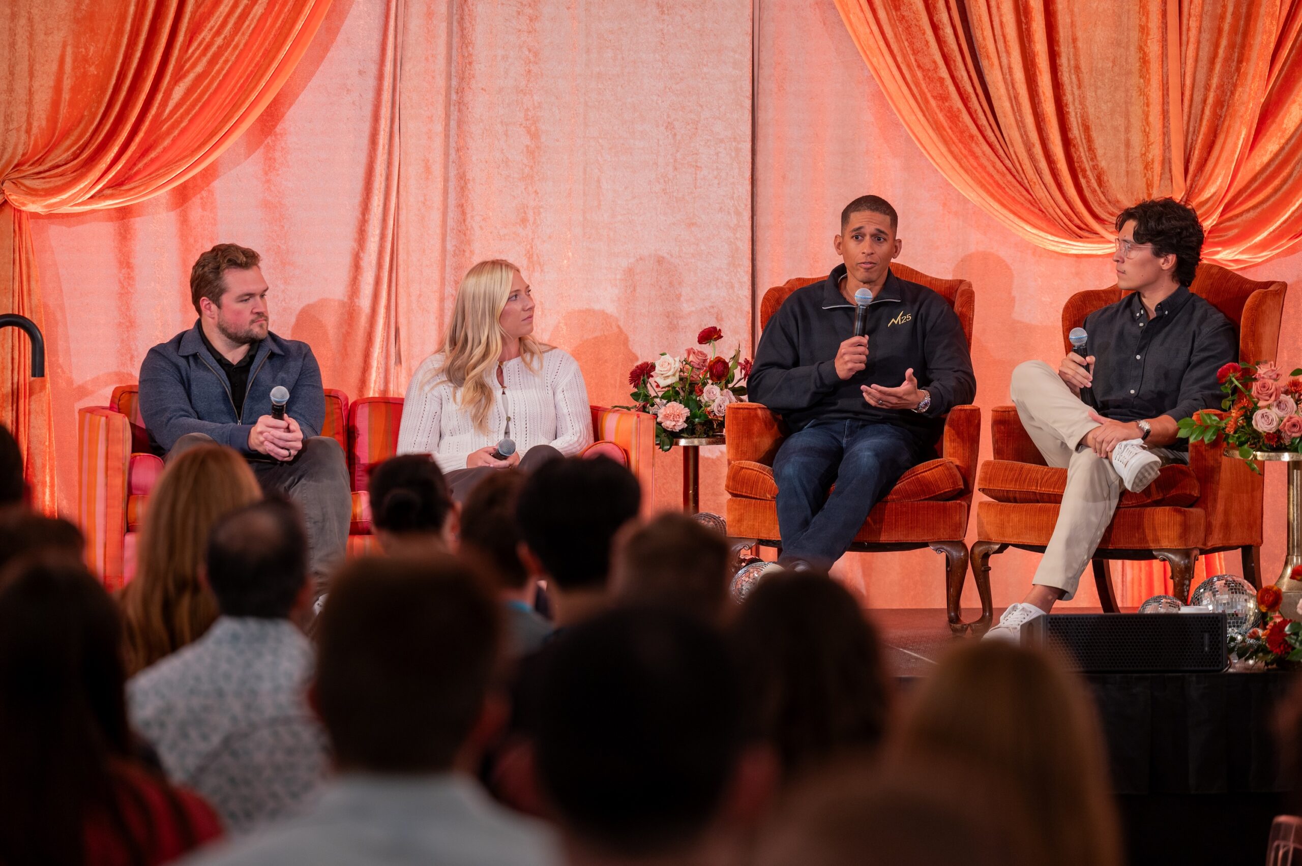 Four panelists seated in armchairs on a stage before an audience, with one speaker holding a microphone and addressing the crowd.
