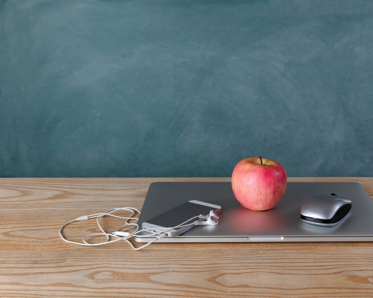 A red apple, smartphone with wired earbuds, and a computer mouse resting on a closed laptop on a wooden desk against a chalkboard.