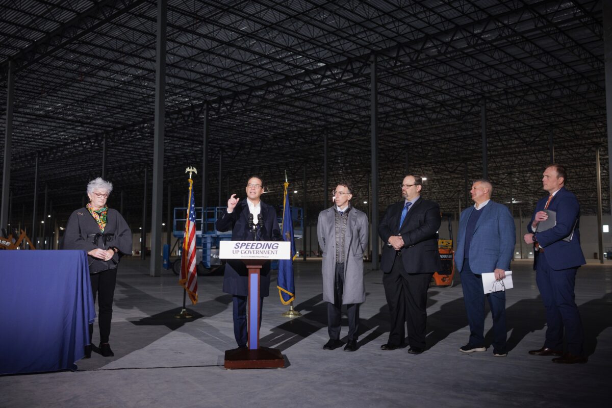 A man speaks at a podium with “SPEEDING UP GOVERNMENT” on it, flanked by four other people in a large, empty industrial building.