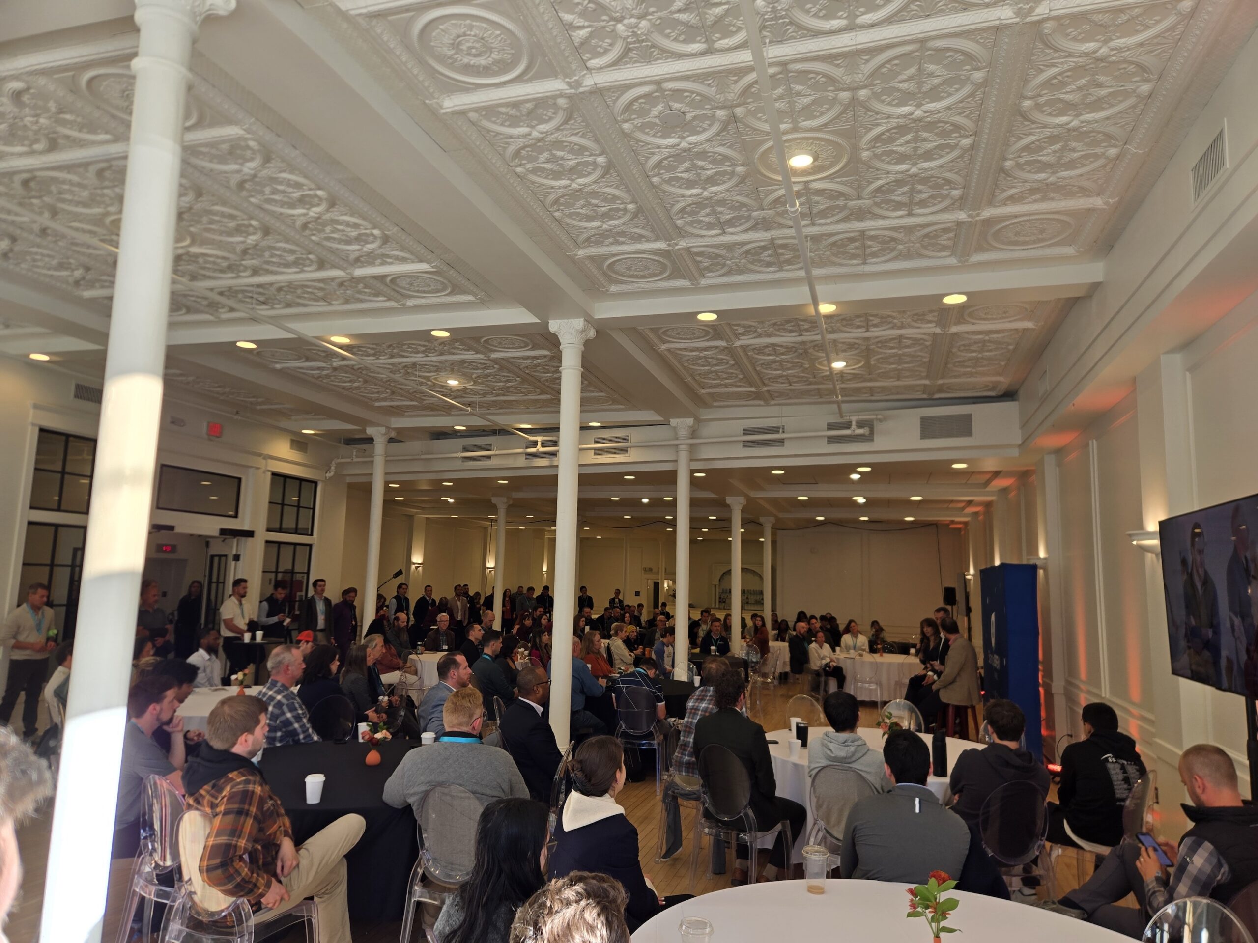 A large audience seated in a hall with an ornate white ceiling listens to a presentation by two speakers on a stage.
