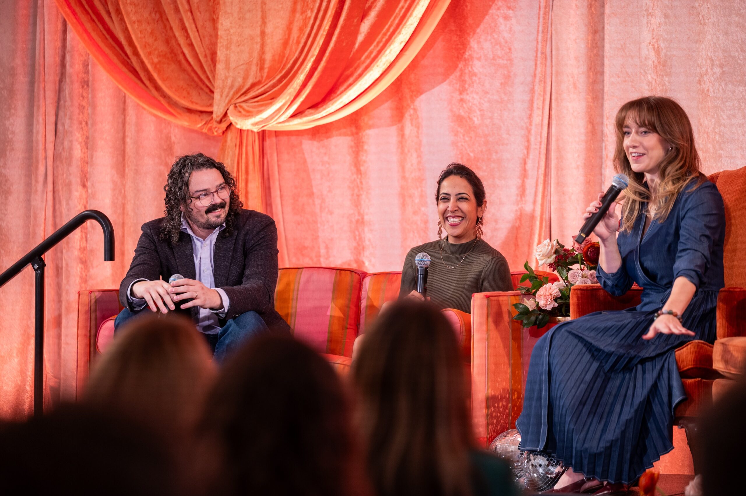 Three panelists sit on stage in armchairs with microphones, speaking to an audience.