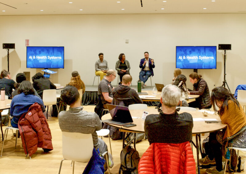 Three people participate in a panel discussion titled “AI & Health Systems” in front of an audience seated at round tables.