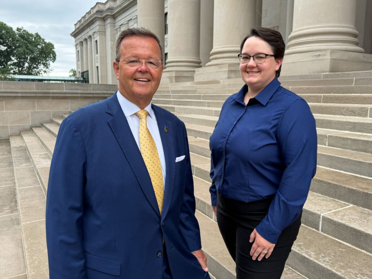 A man in a suit and a woman in a blue shirt stand on the stone steps of a large building.